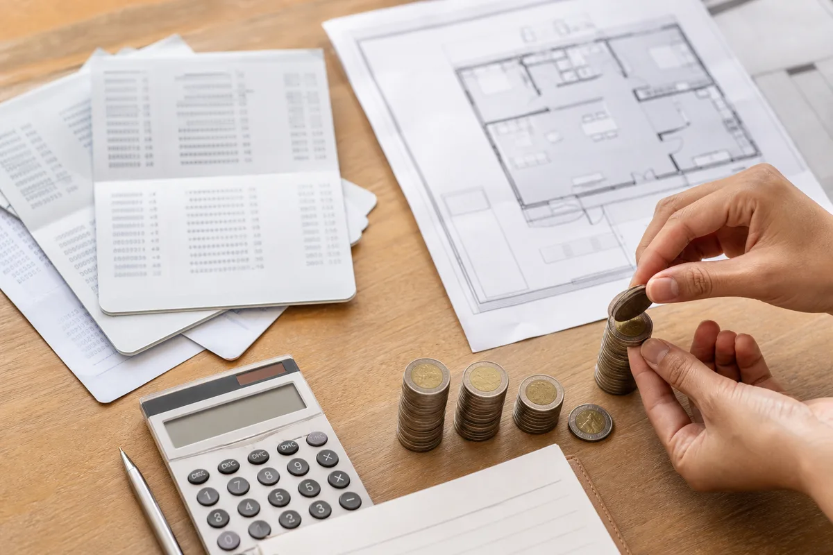 Calculatrice, relevés bancaires et plan de maison sur un bureau, mains posant des pièces d&rsquo;épargne
